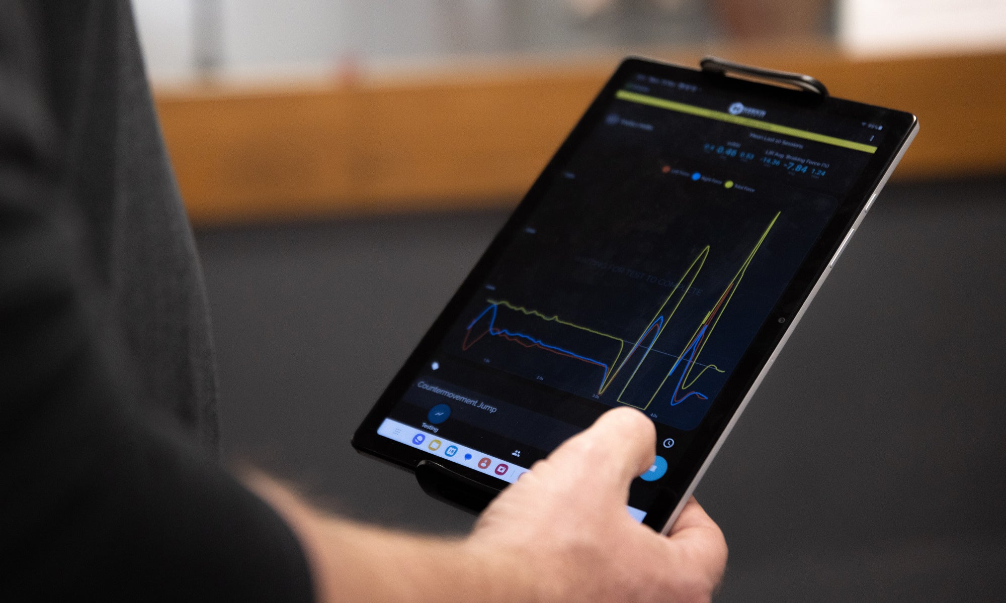 A person holds a tablet displaying a dark-themed graph with several lines and data points, using their right hand to interact with the screen. The background is out of focus.