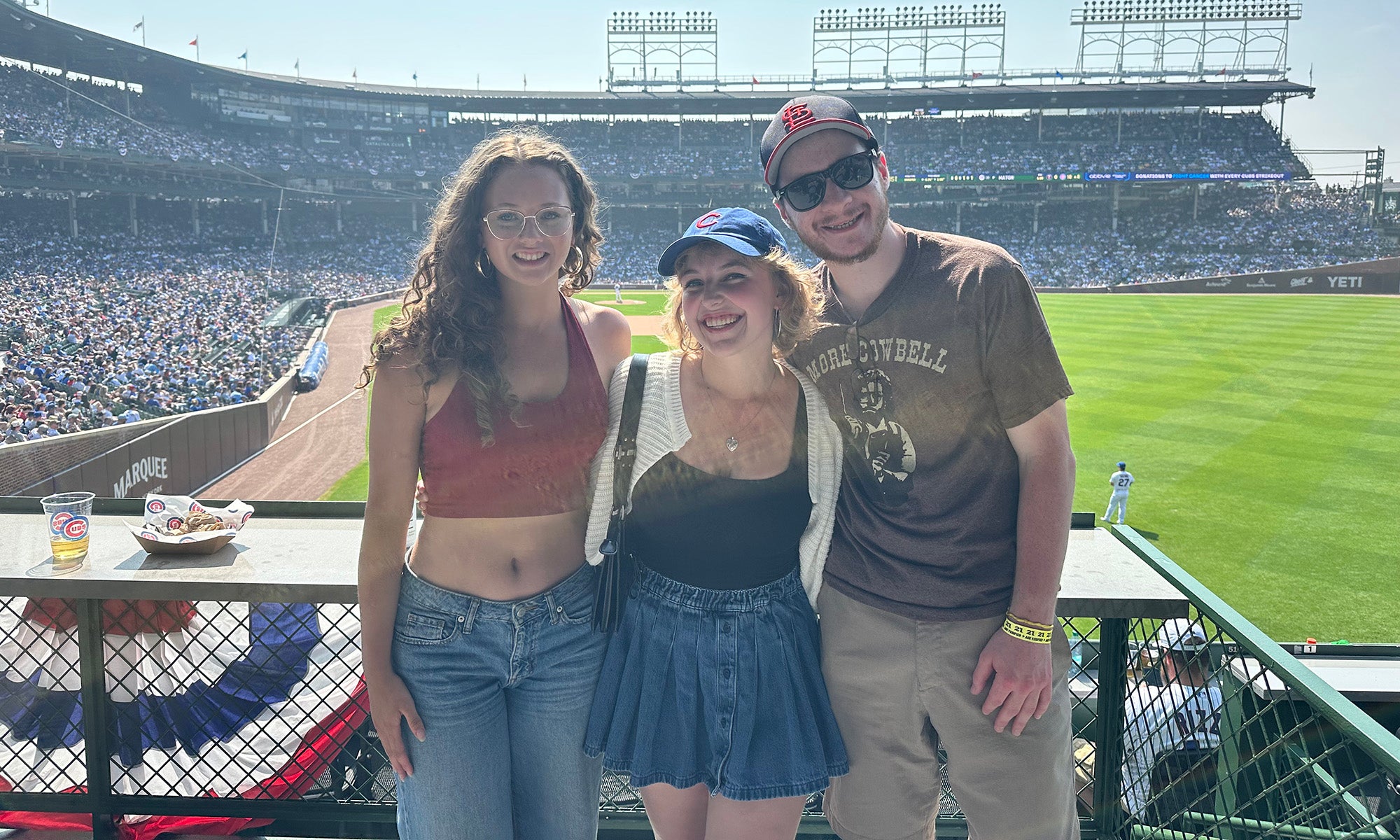 Three people posing and smiling in front of a baseball field, with stands full of spectators and sunlight in the background.