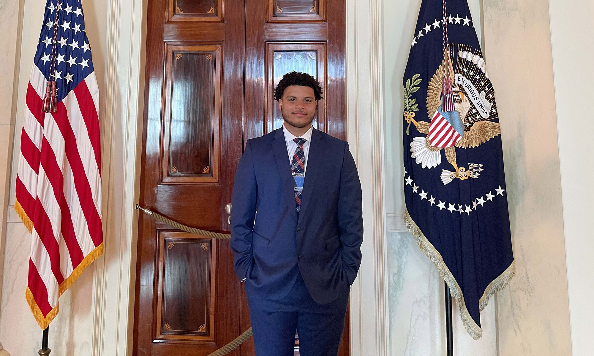 A man in a blue suit stands between the U.S. flag and a presidential flag in front of double wooden doors inside a formal building.