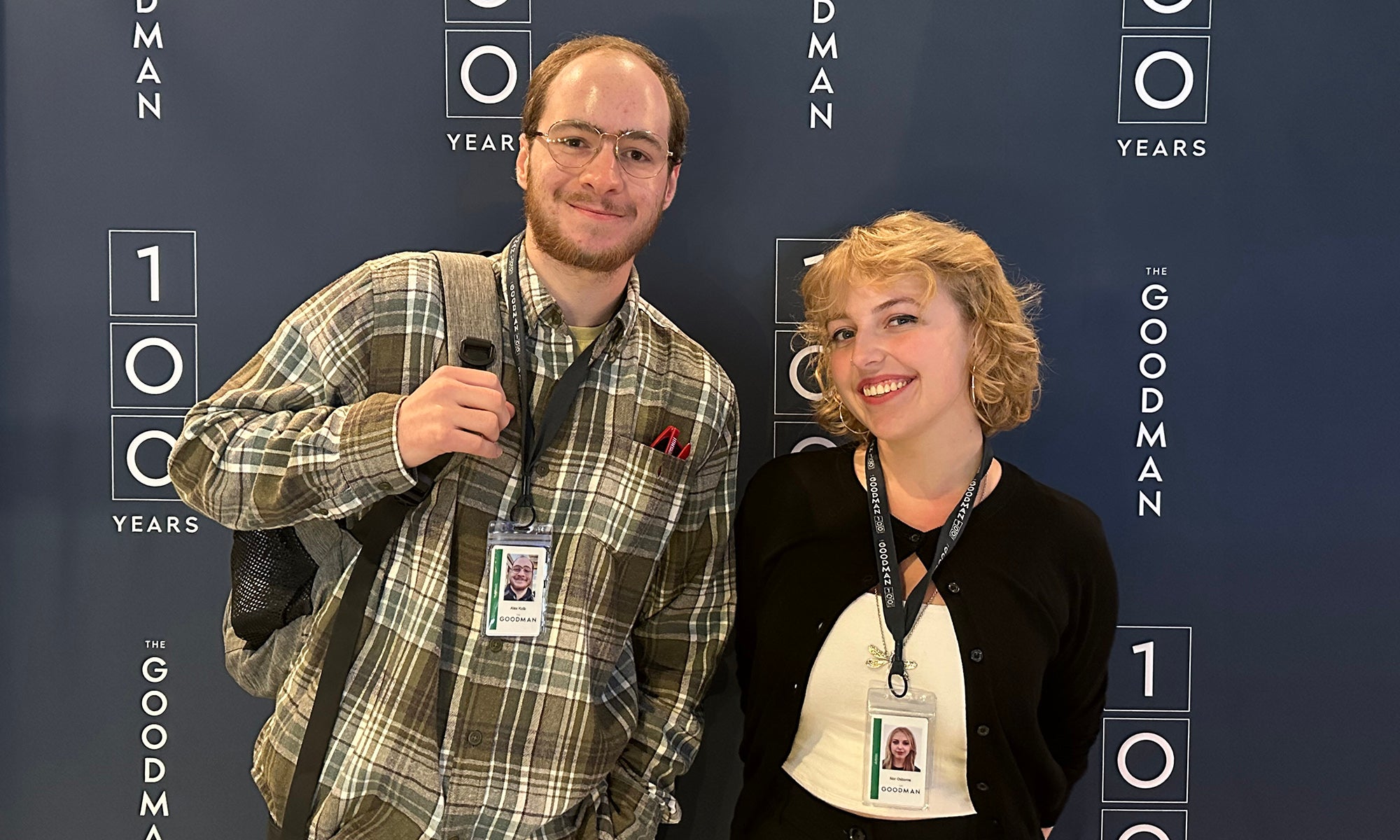 Two people with ID badges pose and smile in front of a blue backdrop with 100 Years Goodman printed repeatedly in white letters.