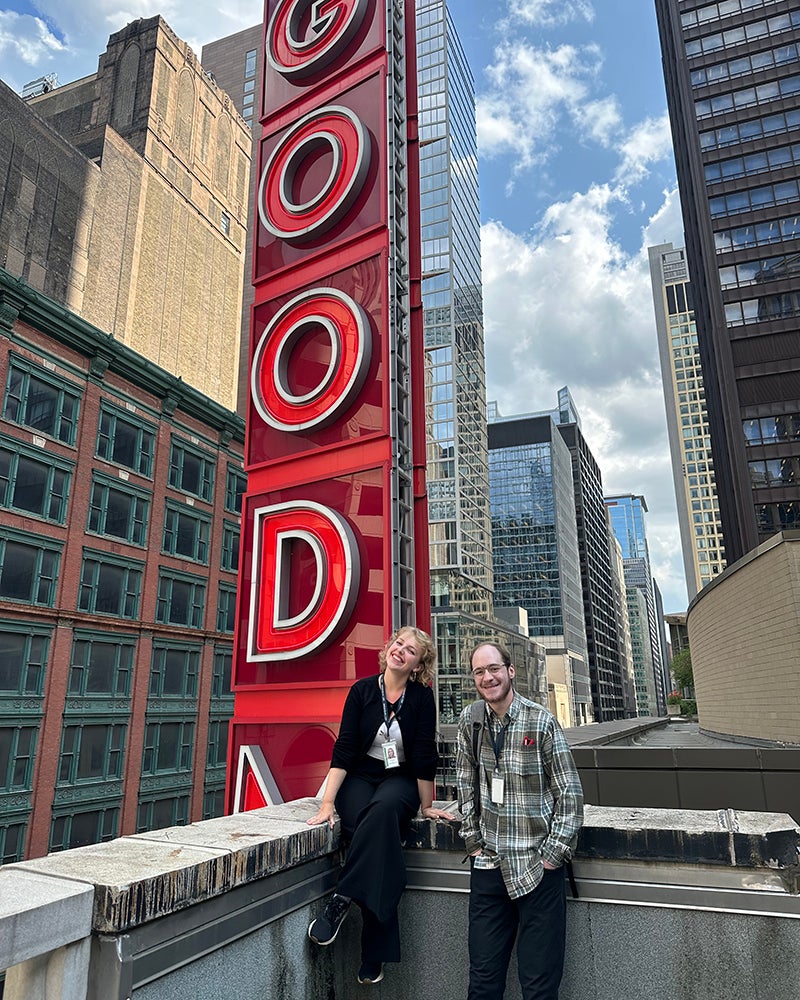 Two people stand on a rooftop in front of a large vertical red sign that reads GOOD with city buildings and a partly cloudy sky in the background.