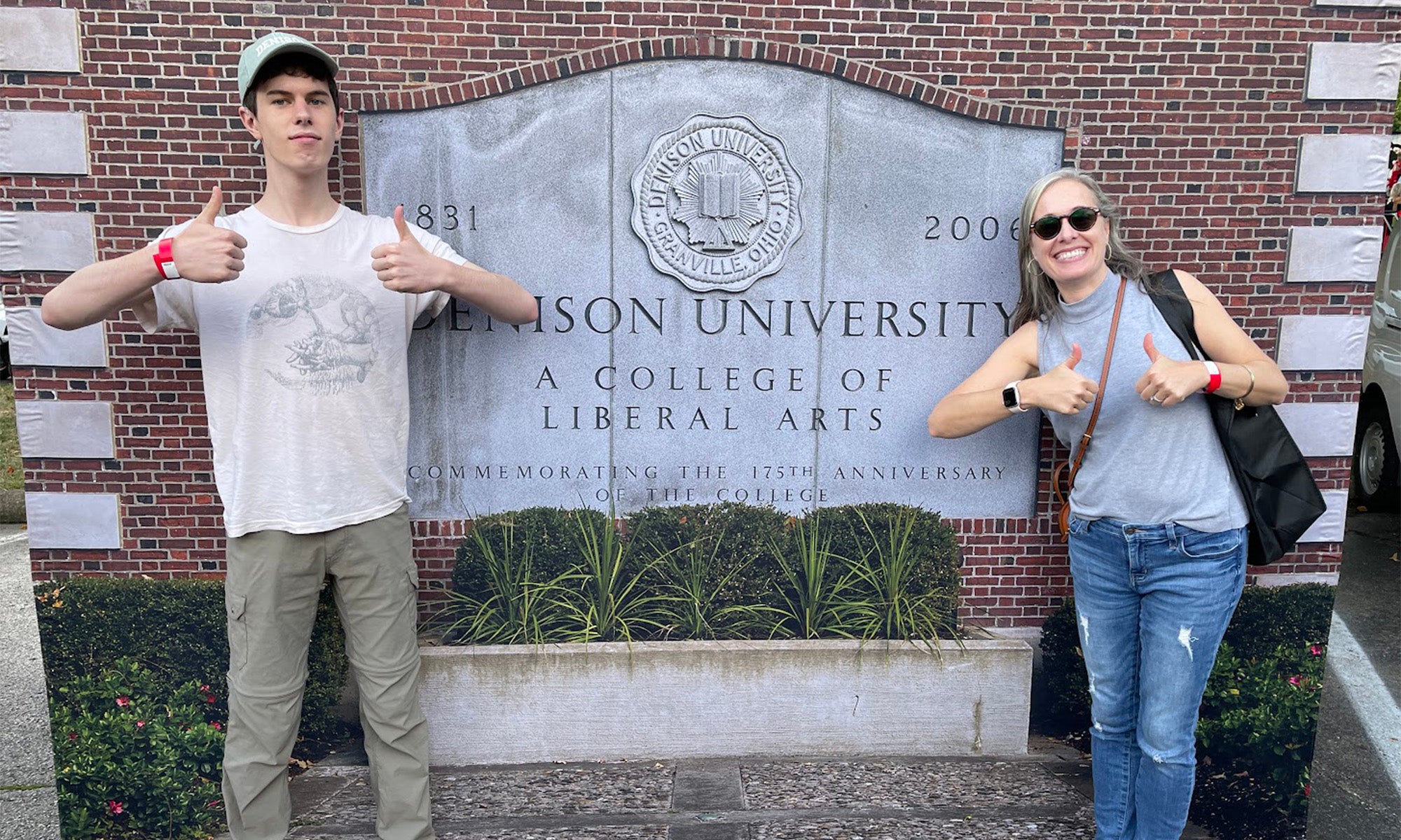 Two people stand in front of a brick wall with a sign reading “Denison University: A College of Liberal Arts.” Both are smiling and giving thumbs-up gestures.