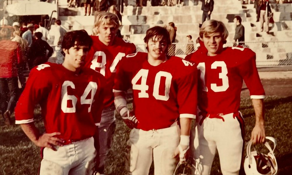 Four football players in red jerseys with numbers 64, 84, 40, and 13 stand on a field; bleachers and spectators are visible in the background.