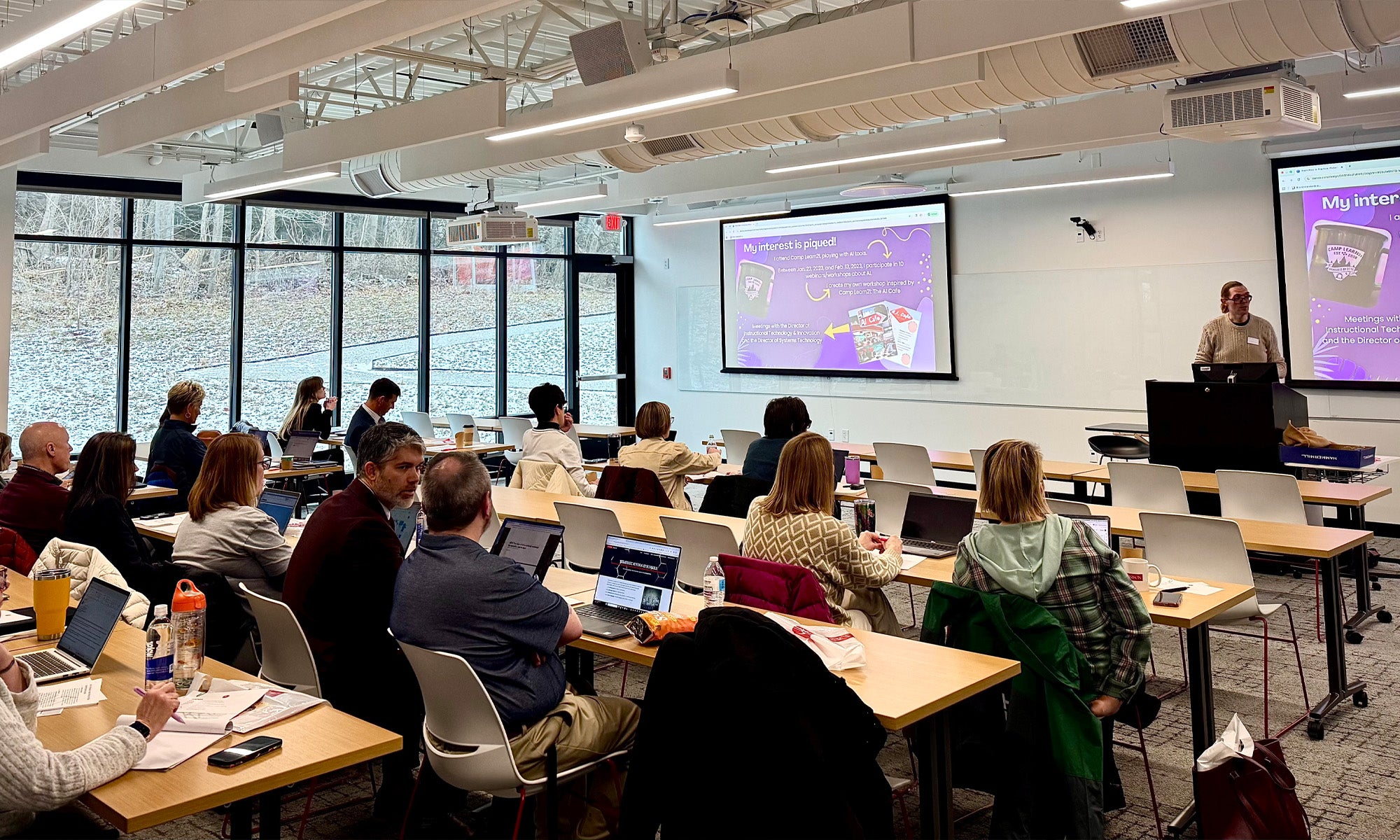A classroom with adults seated at tables using laptops, listening to a presenter standing by screens displaying a colorful slide presentation.