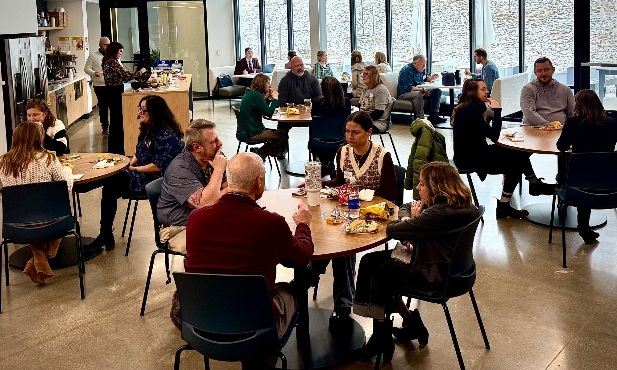 Conference participants seated at round tables in an modern office cafeteria space; a large wall of windows in the background.