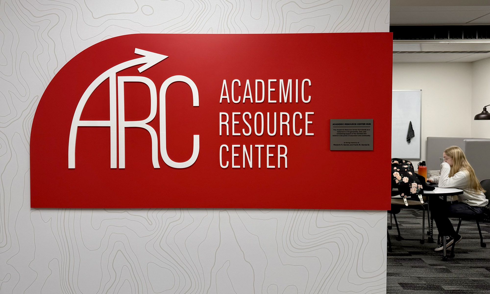 Red sign reading ARC Academic Resource Center mounted on a wall; in the background, a person sits at a desk in a study space.