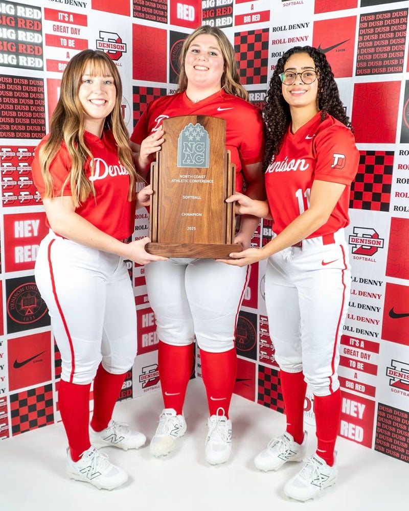 Three softball players in red and white Denison uniforms stand together, holding a large NCAC championship trophy in front of a red and black Denison-themed backdrop.