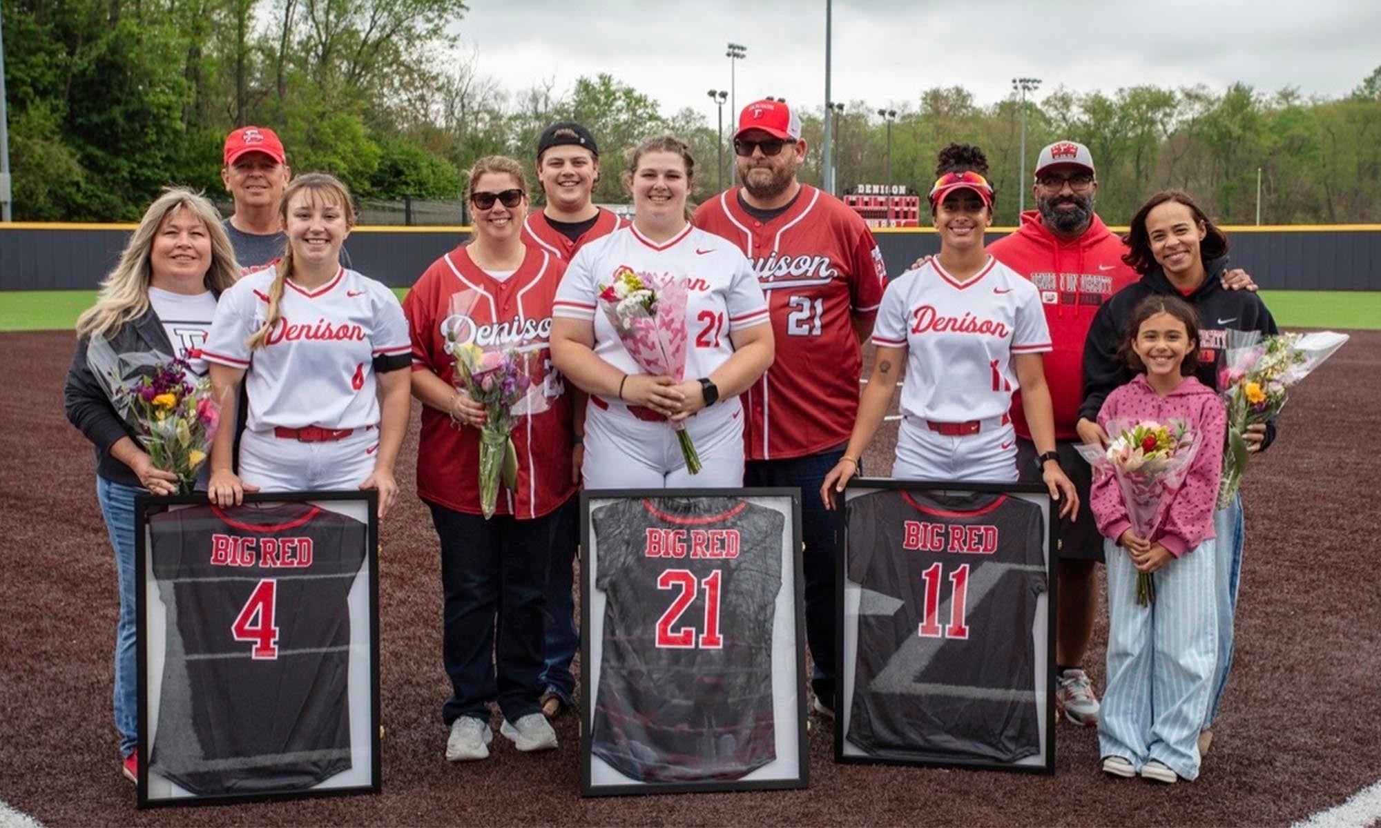 A group of people poses on a softball field with framed jerseys and bouquets, celebrating senior players wearing Denison uniforms numbered 4, 21, and 11.