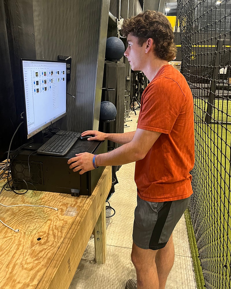 A person in a red shirt and shorts stands at a wooden table, using a computer in an indoor sports or training facility.