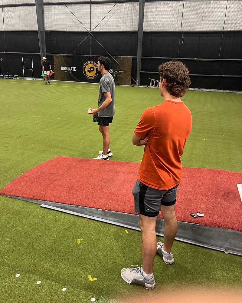 Two men stand on an indoor turf field, one on a red mat and the other nearby, both wearing athletic clothing.