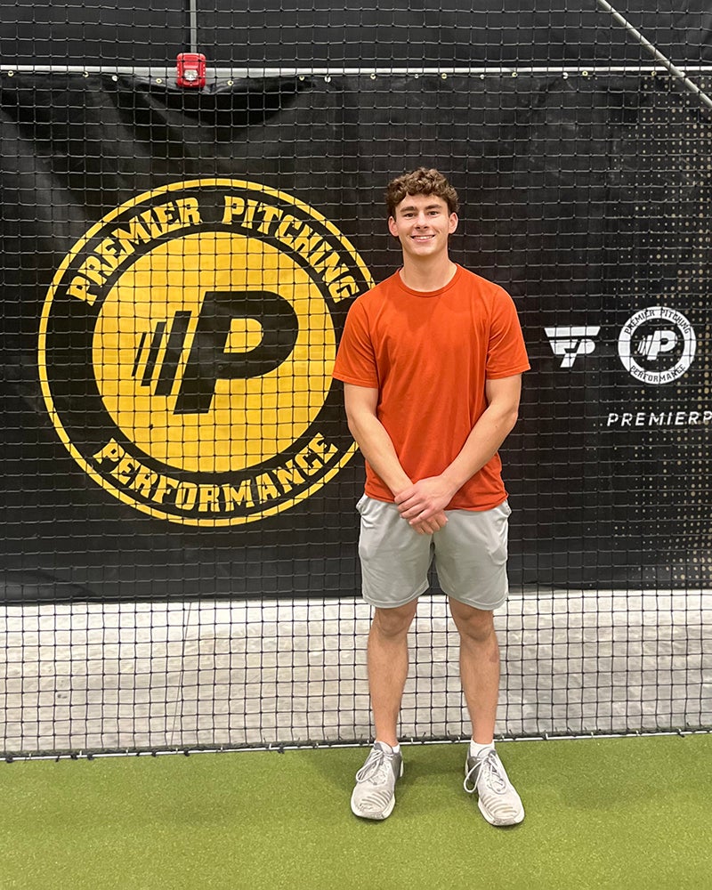 Young man in an orange shirt and gray shorts stands indoors in front of a Premier Pitching Performance banner on a netted wall.