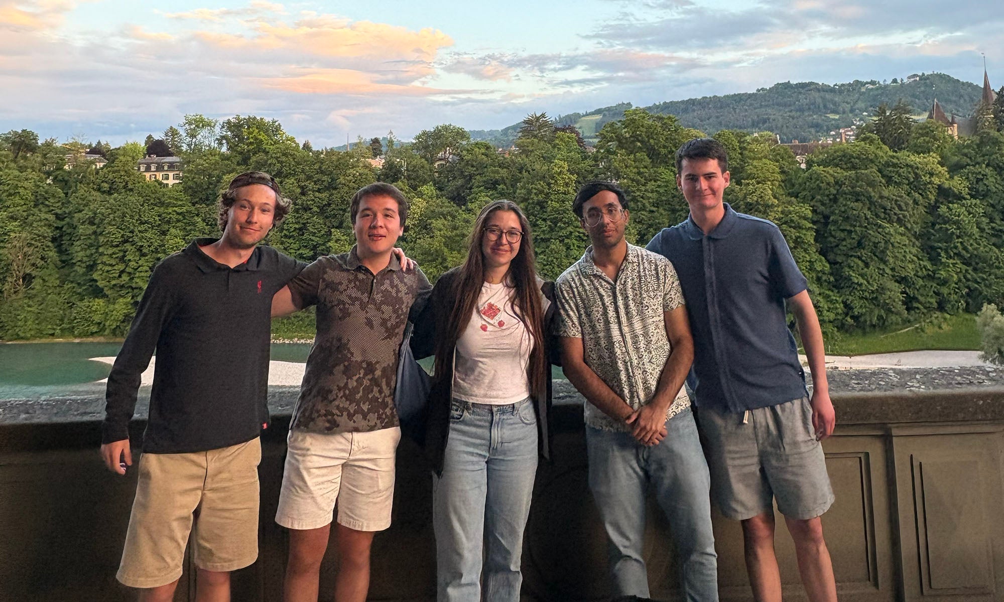 A group of people stand by a stone wall overlooking a verdant mountain range at sunset.
