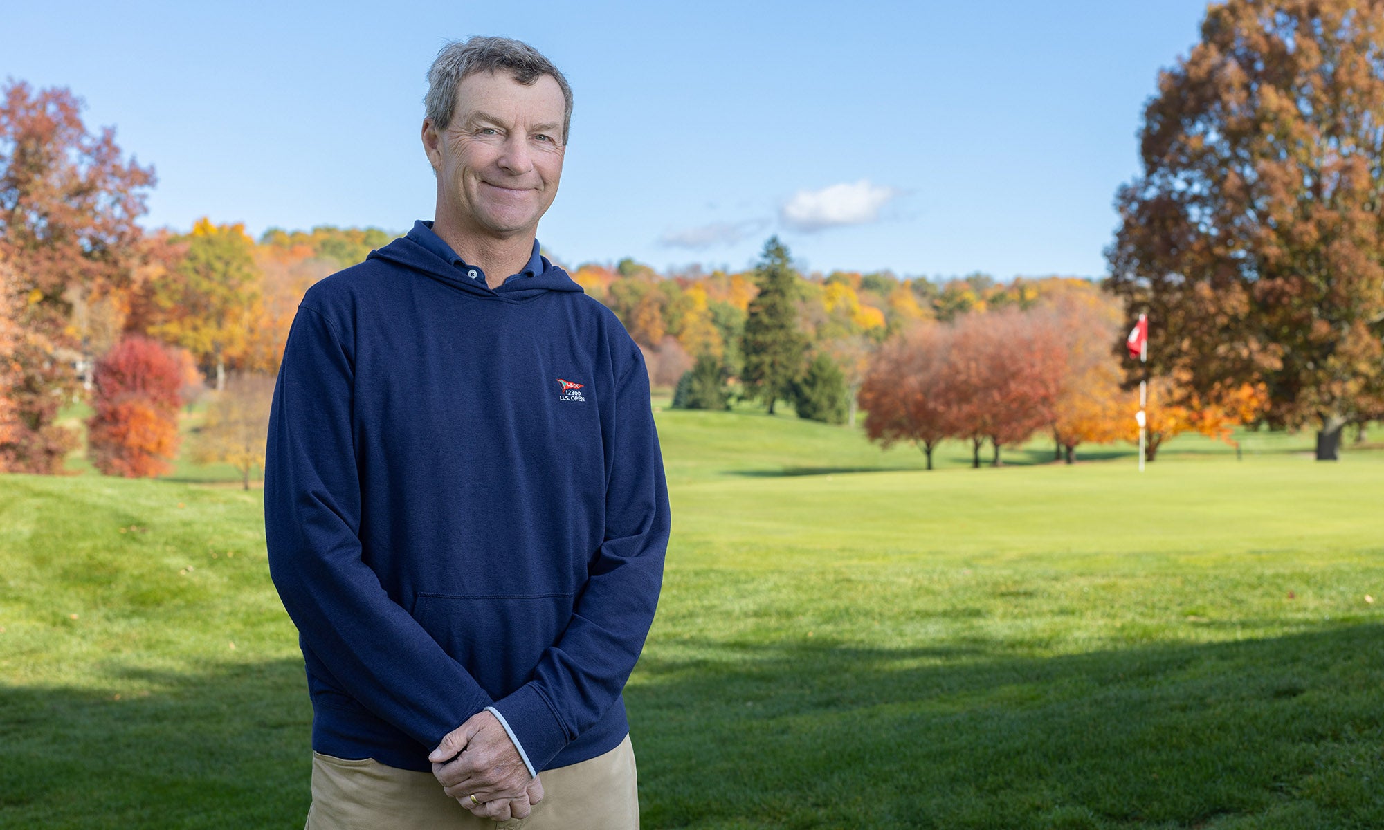 A person stands on a grassy field with autumn trees and a partly cloudy sky in the background.
