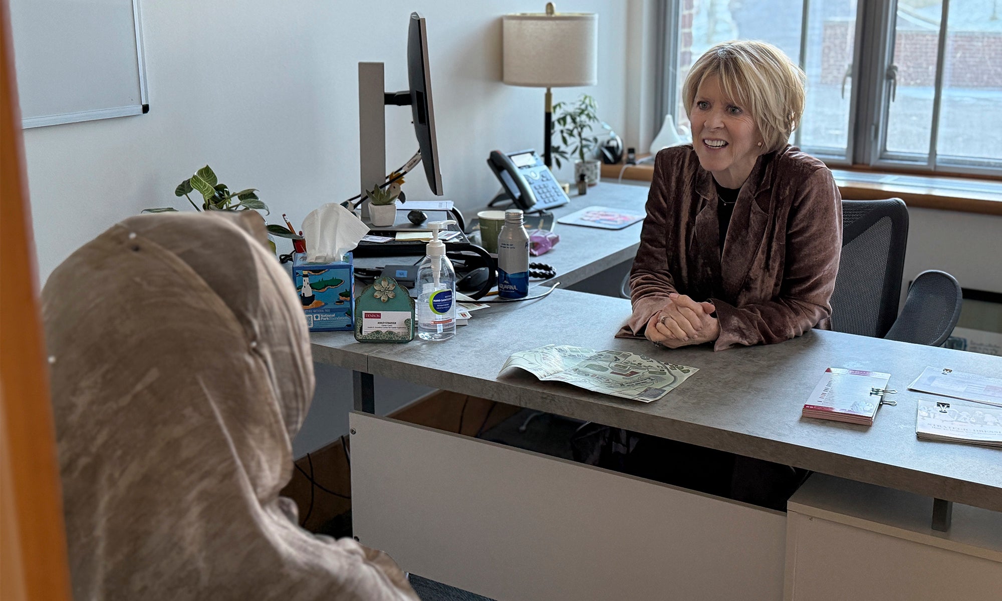 A woman sitting by a window at a desk in a business office talking to an person seated across from them.