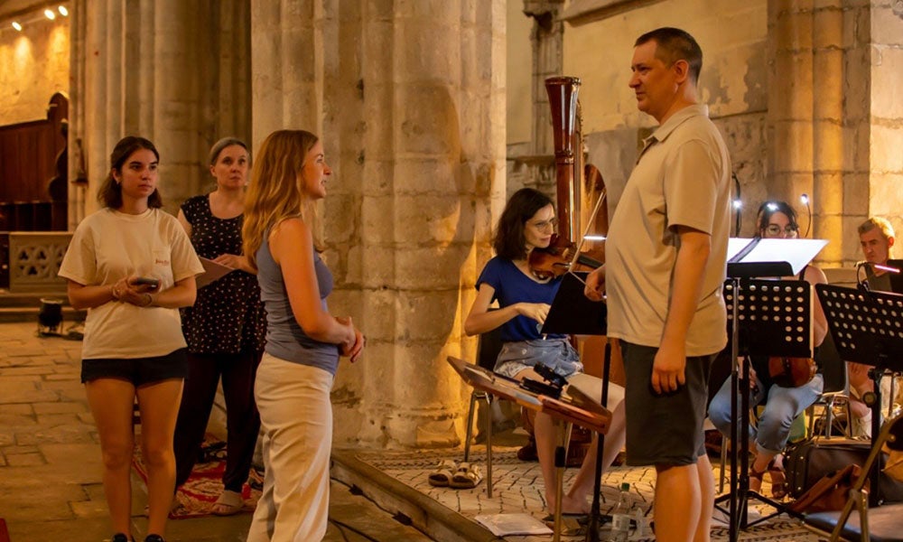 Several people stand and converse near musicians seated with instruments inside a stone building, possibly during a rehearsal or informal performance.