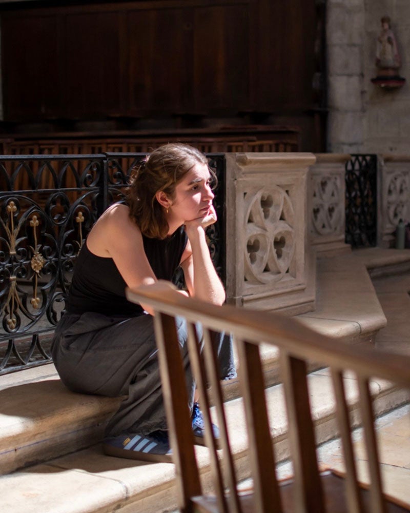 A woman sits on stone steps indoors, resting her chin on her hand and looking to the side in natural light.