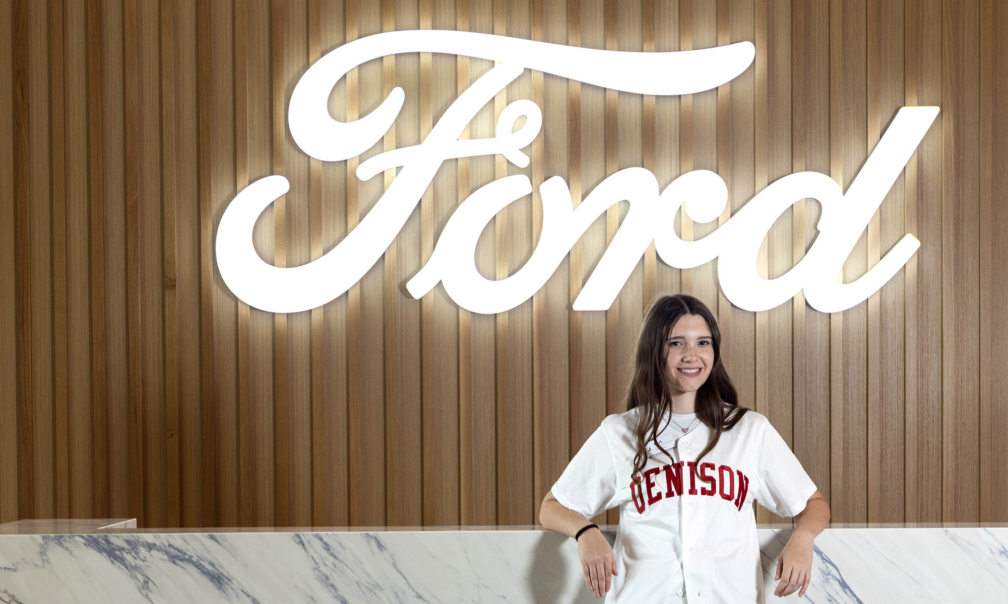 A person wearing a Denison shirt stands in front of a large illuminated Ford logo on a wooden wall.