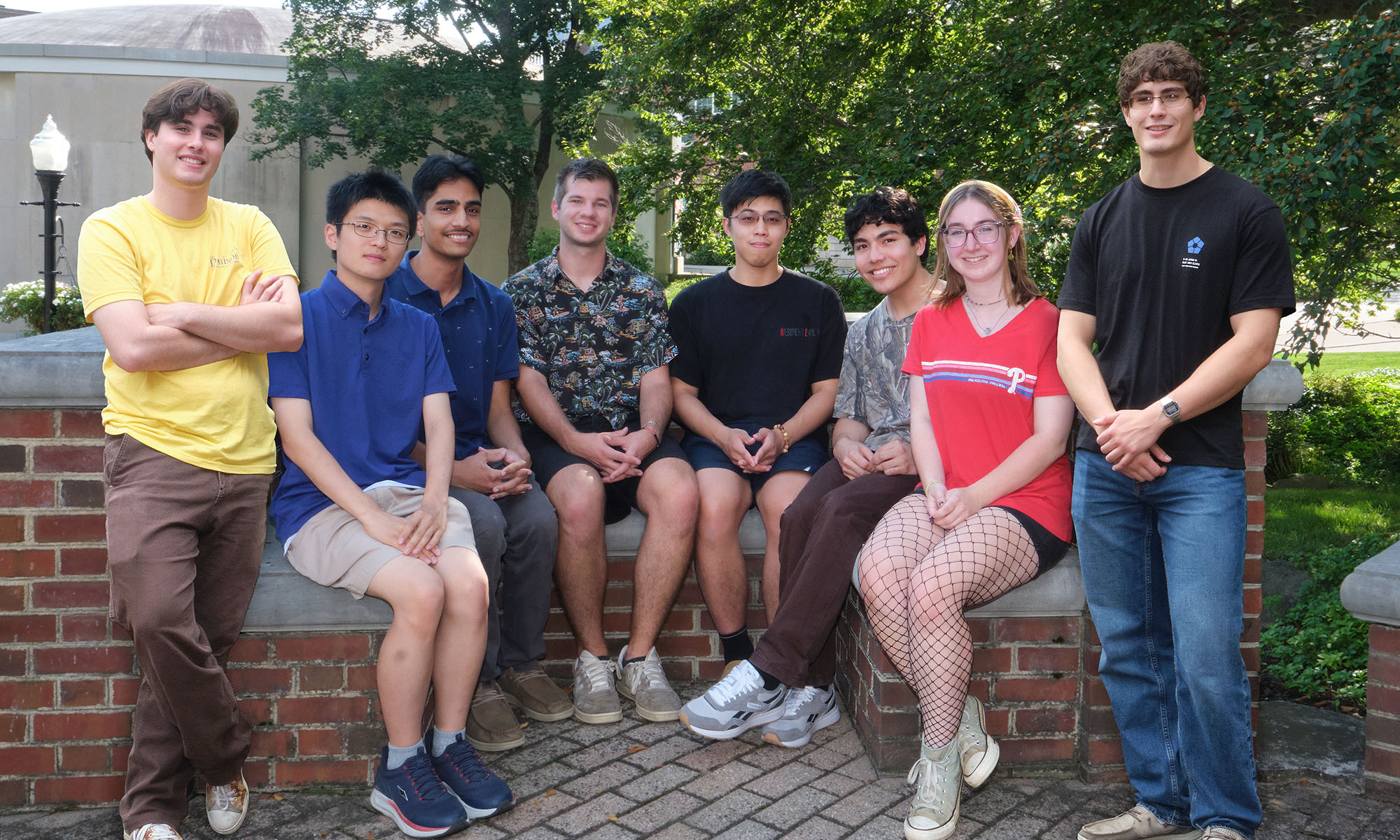 Eight young adults pose together outdoors on a brick structure, with trees and greenery in the background.