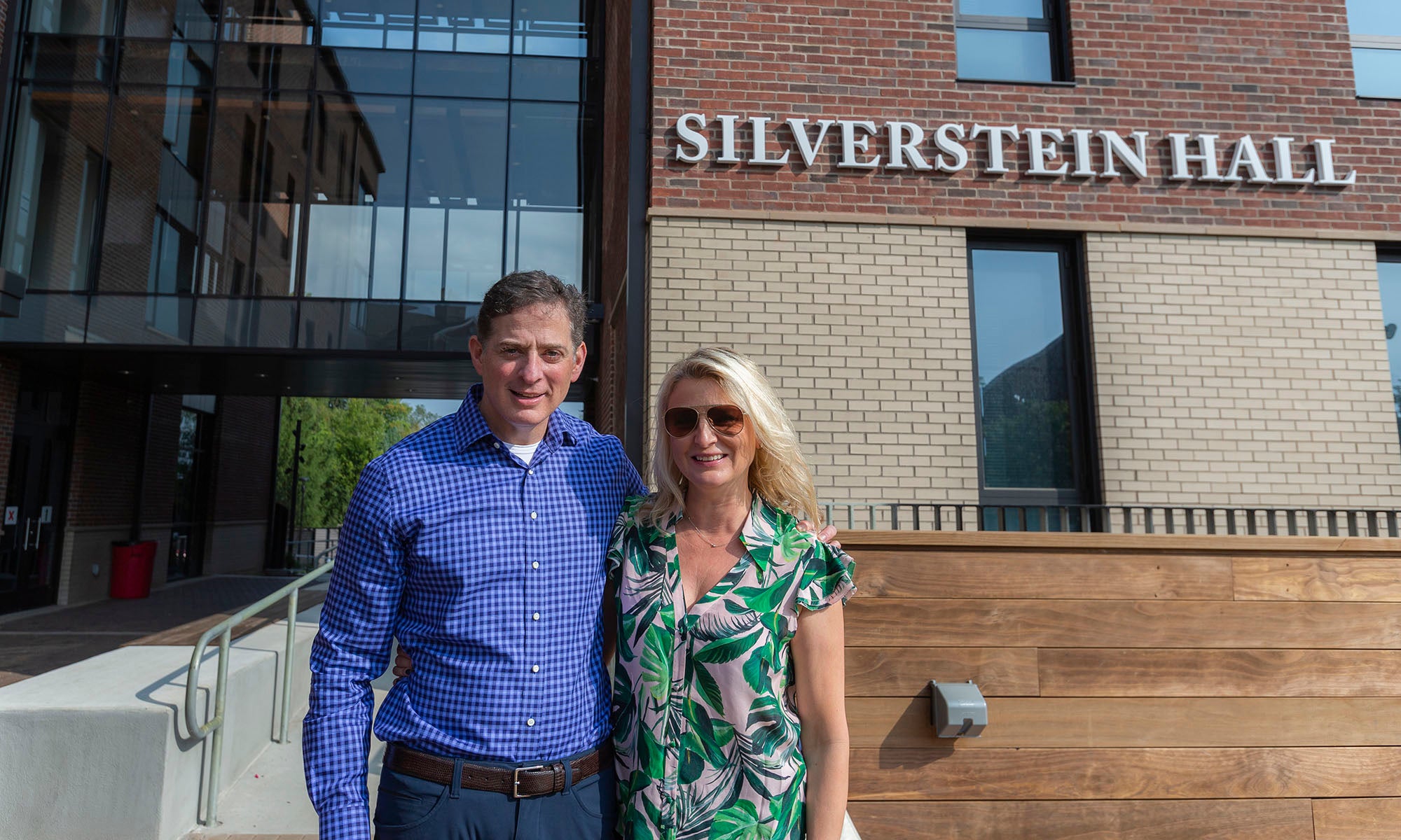A man and a woman stand together outside a building with large windows and a sign that reads Silverstein Hall.