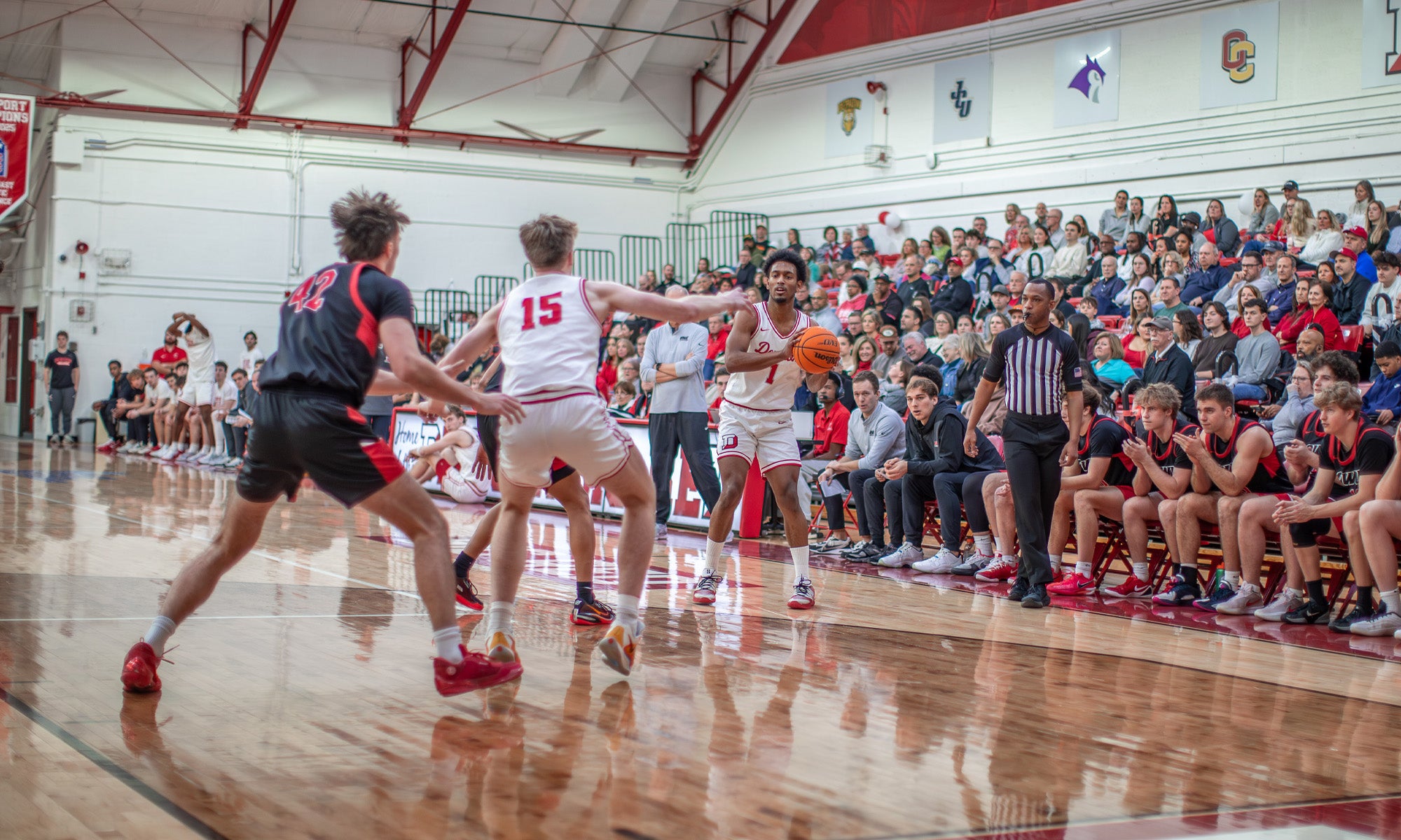 Basketball players play in a large gymnasium; bleachers packed with fans cheering the background.
