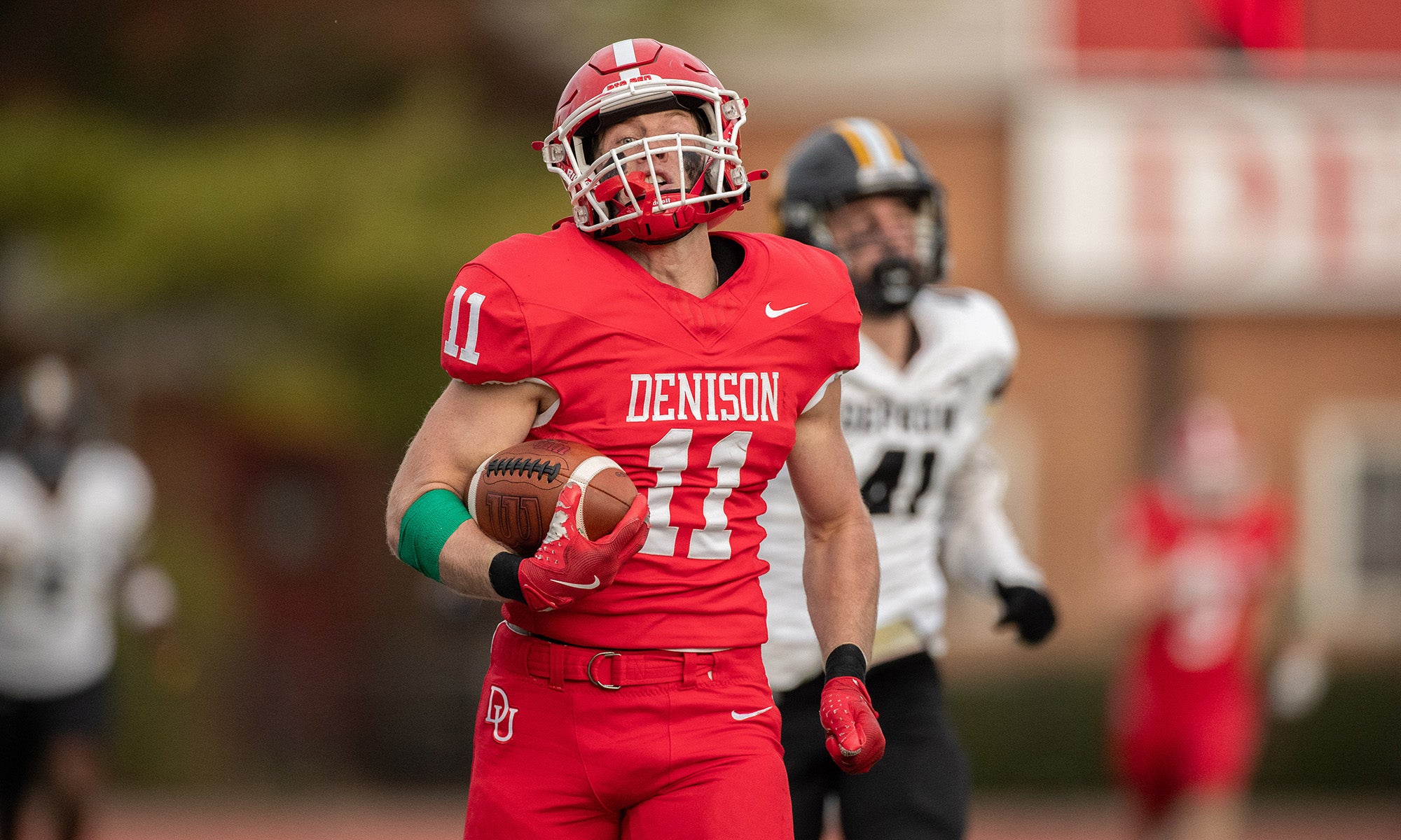 A football player in a red Denison uniform runs with the ball on the field, with a defender in a white uniform in the background.