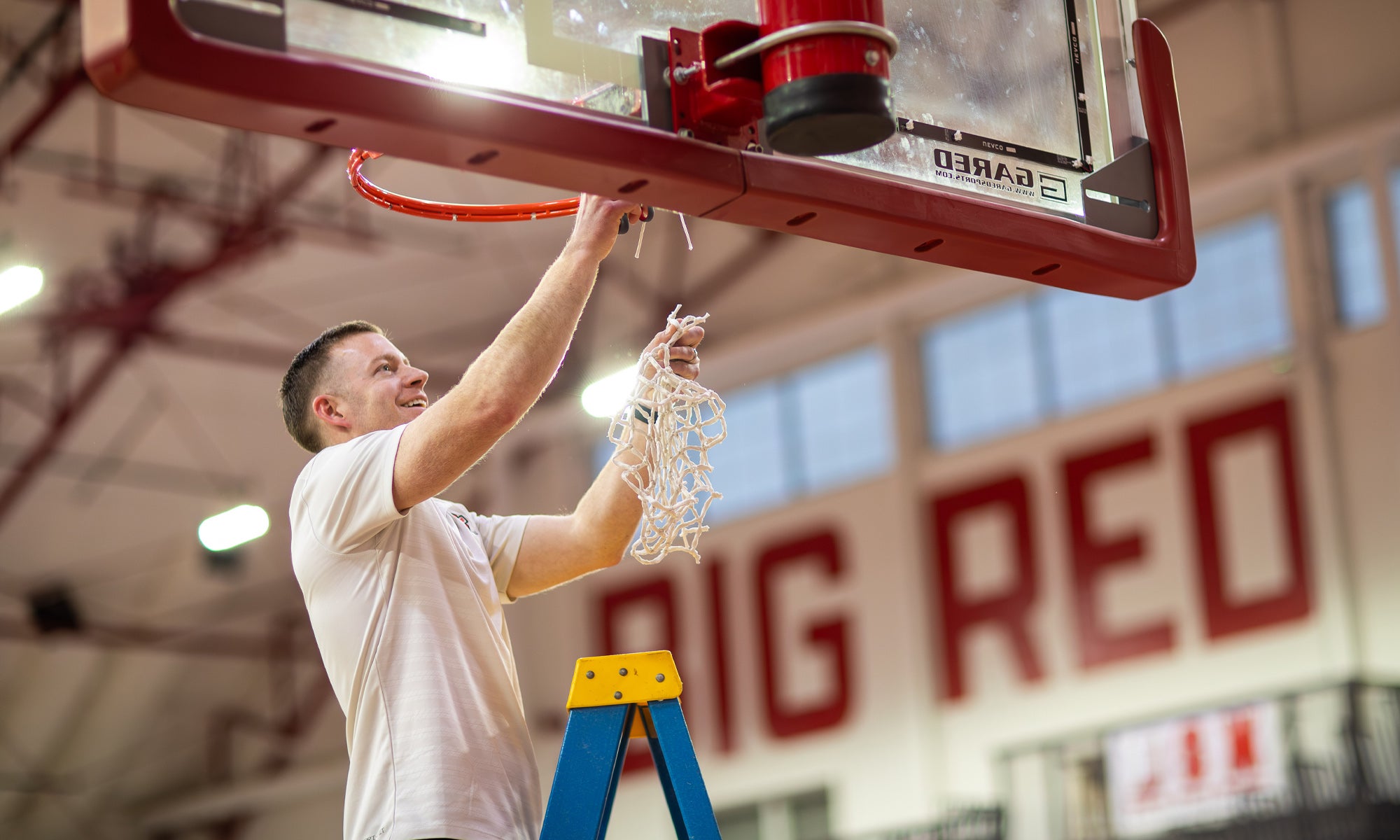 A man in a light tee shirt stands on a blue latter and cuts down a basketball net from a gymnasium backboard.