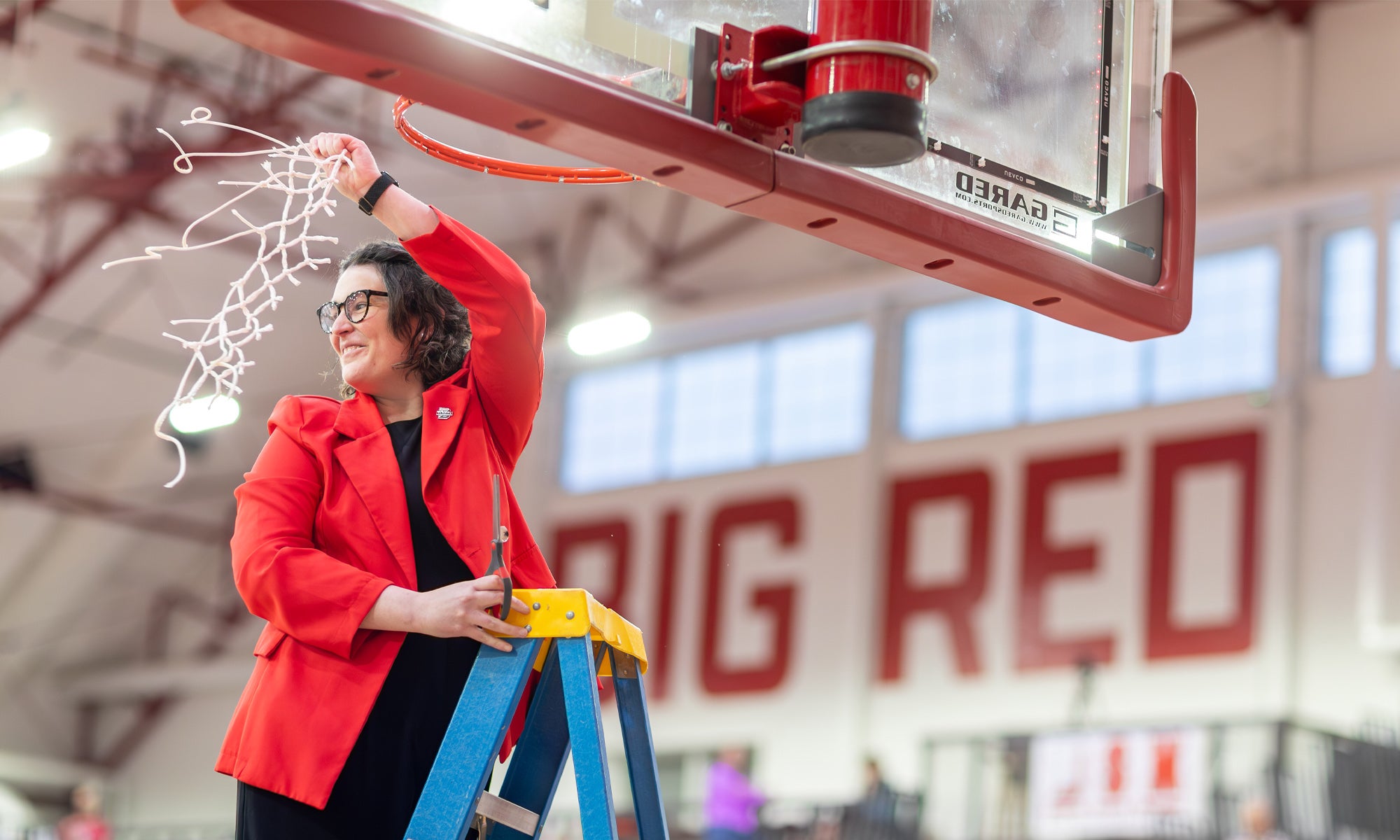 A woman in a red jacket and black pants stands on a blue ladder and cuts down a basketball net from a gymnasium backboard.
