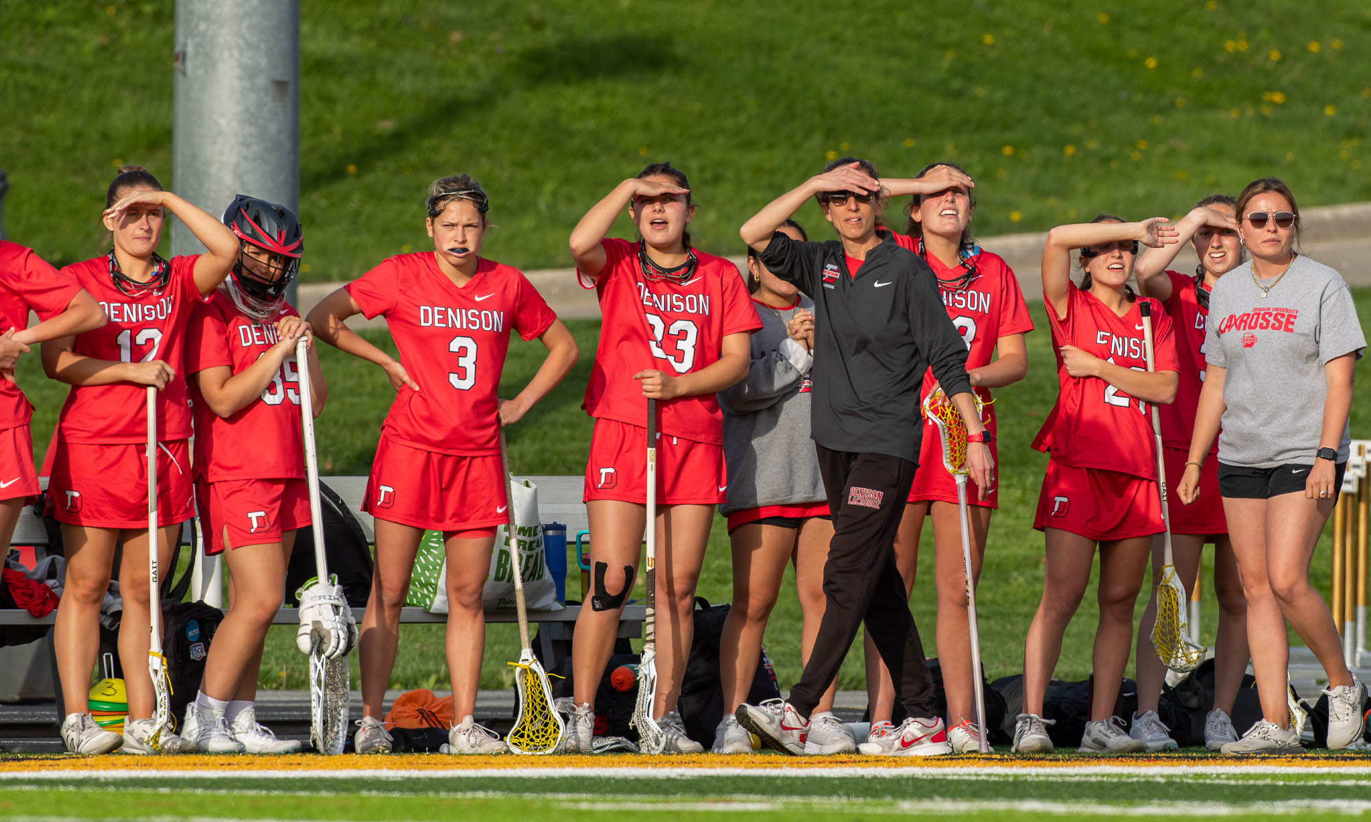 A group of women's lacrosse players and their coach watch their teammates from the sideline.