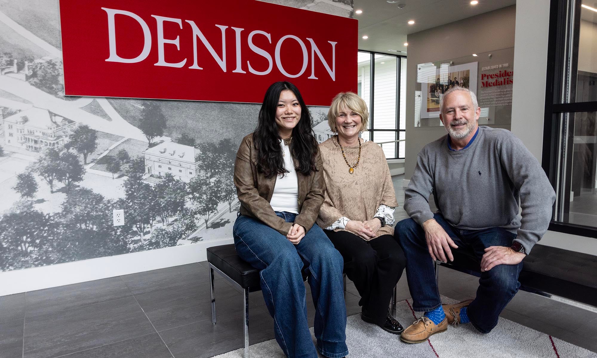 Three people, including Provost Kim Coplin, sit on a bench indoors in front of a large red Denison banner. They are smiling and posing for the photo in a modern, well-lit space.