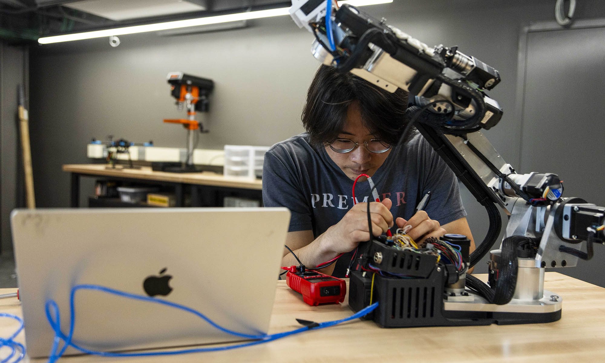 A person works on wiring or repairing a robotic arm at a desk with a laptop and tools in a workshop setting.