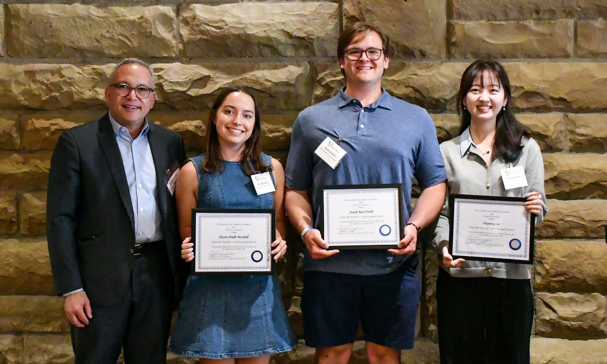 Four people stand in front of a stone wall; three hold certificates and smile, while the fourth person, in a suit, stands beside them.