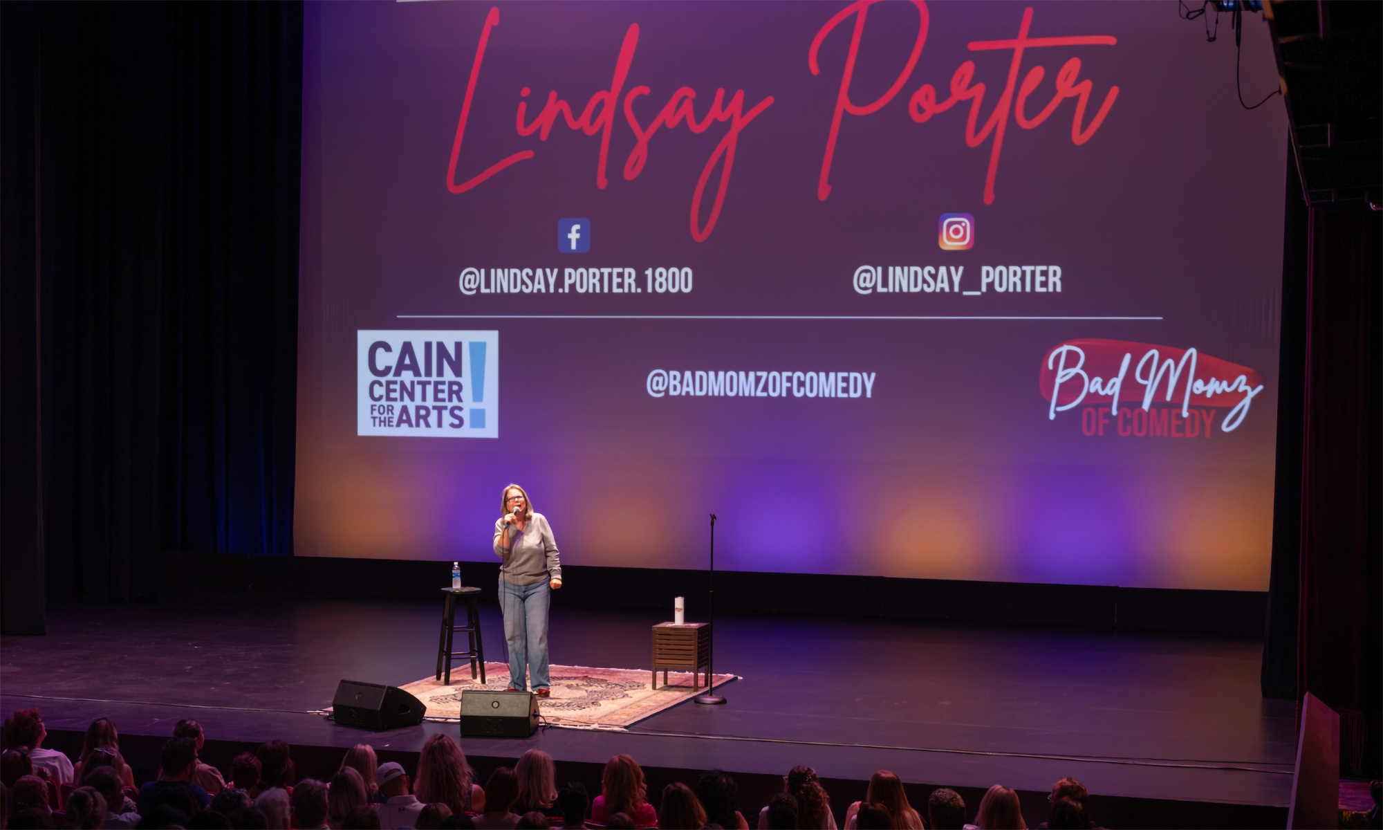 Comedian Lindsay Porter performing on a stage in front of a screen displaying their social media handles and show branding.