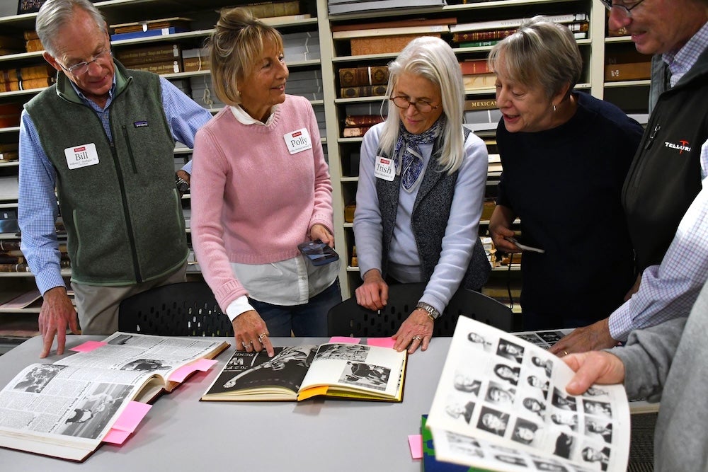 Five adults stand around a table in a library or archive, examining and discussing several open photo albums and yearbooks. Shelves filled with books and files are visible in the background.