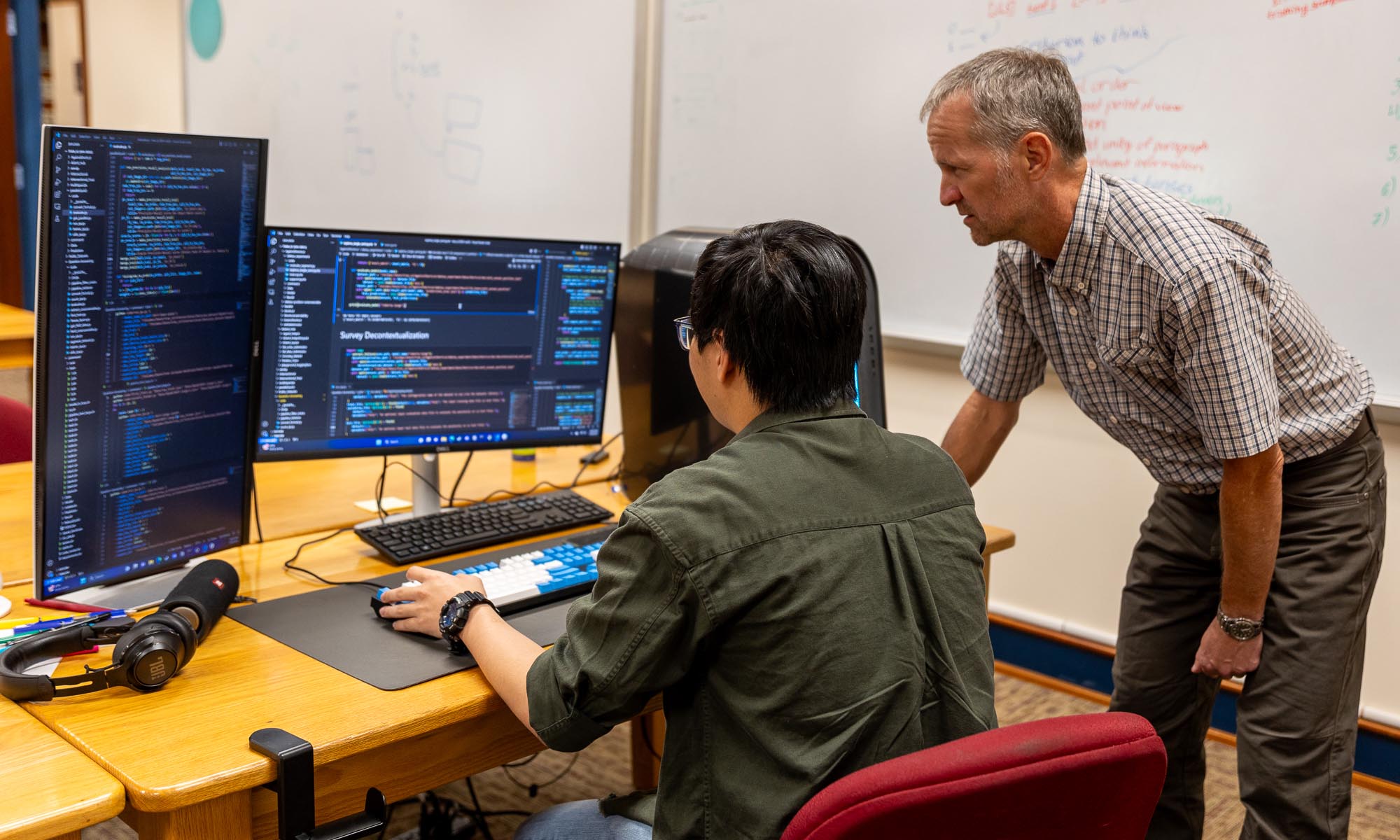 Two people review code on multiple computer monitors in an office; one is seated and typing while the other stands beside, looking at the screens.