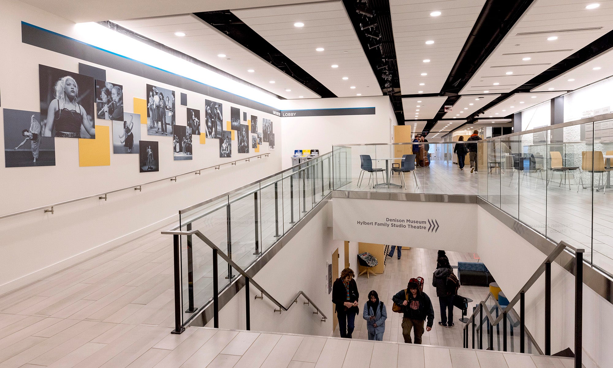 A modern interior hallway with people walking up stairs, wall art featuring musicians on the left, and seating areas on the upper level.