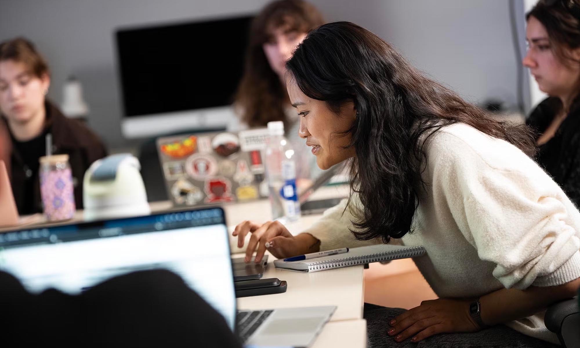 Several people sit around a table with laptops and notebooks, engaged in discussion in a modern office setting.
