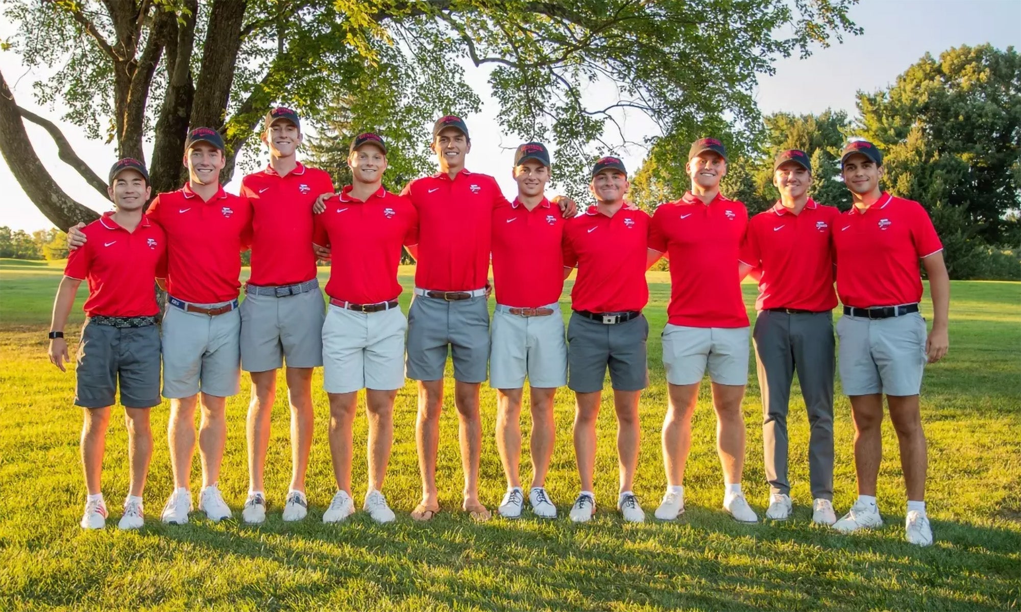Ten young men in matching red shirts and light shorts stand side by side on a grassy area with trees in the background, posing for a group photo outdoors.