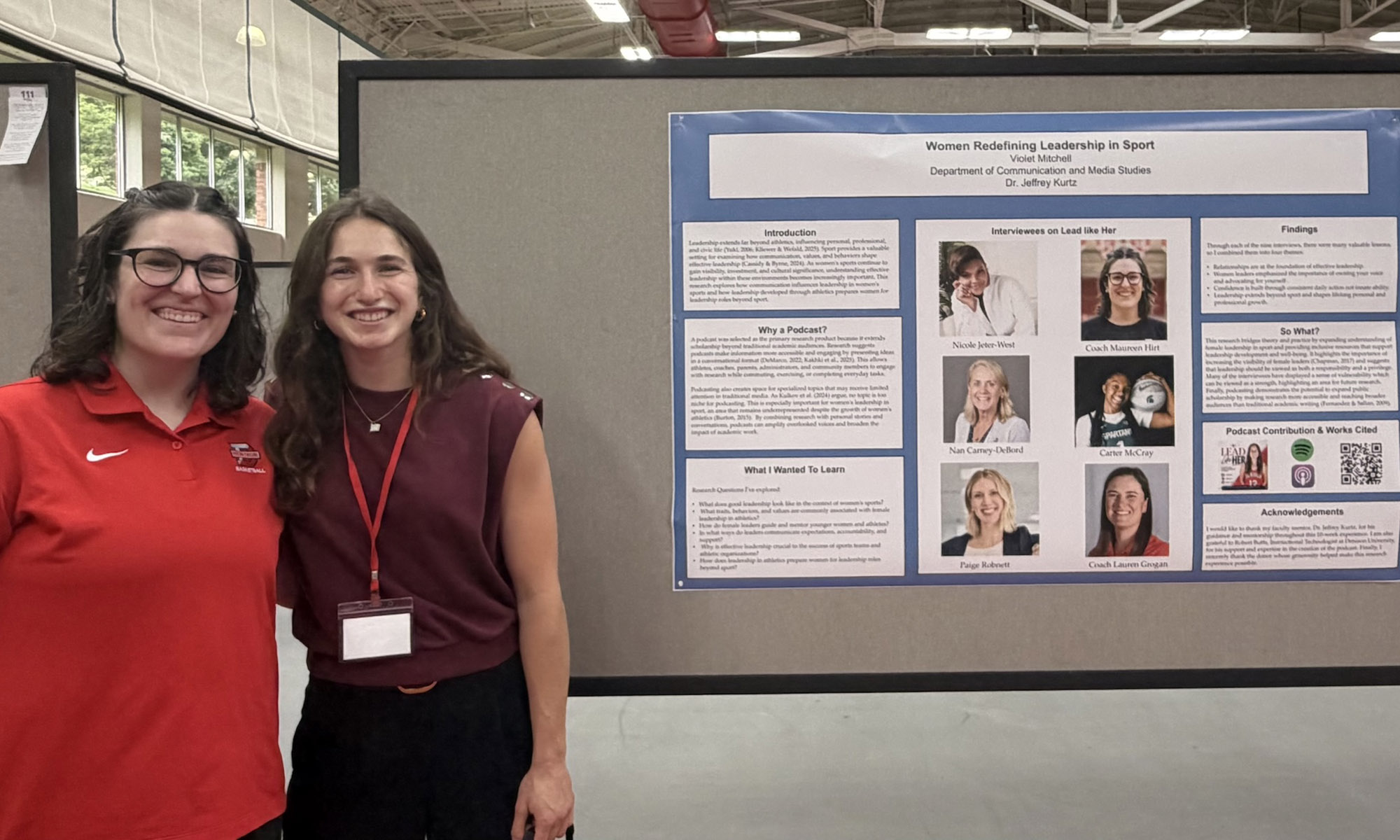 Two women stand smiling in front of a large research poster displayed on a bulletin board in a well-lit indoor setting.
