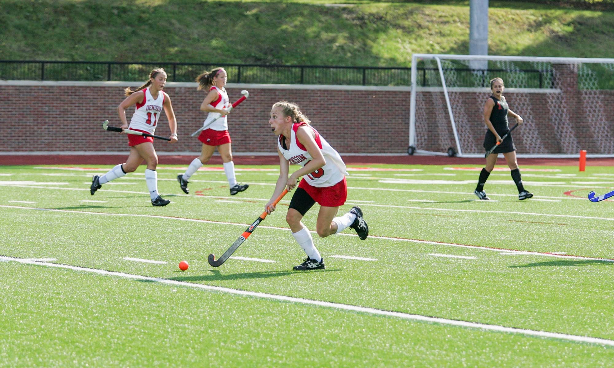 A group of female field hockey players in red and white uniforms play on a green turf field. One player is dribbling the ball while others run behind her. A goal and net are visible in the background.