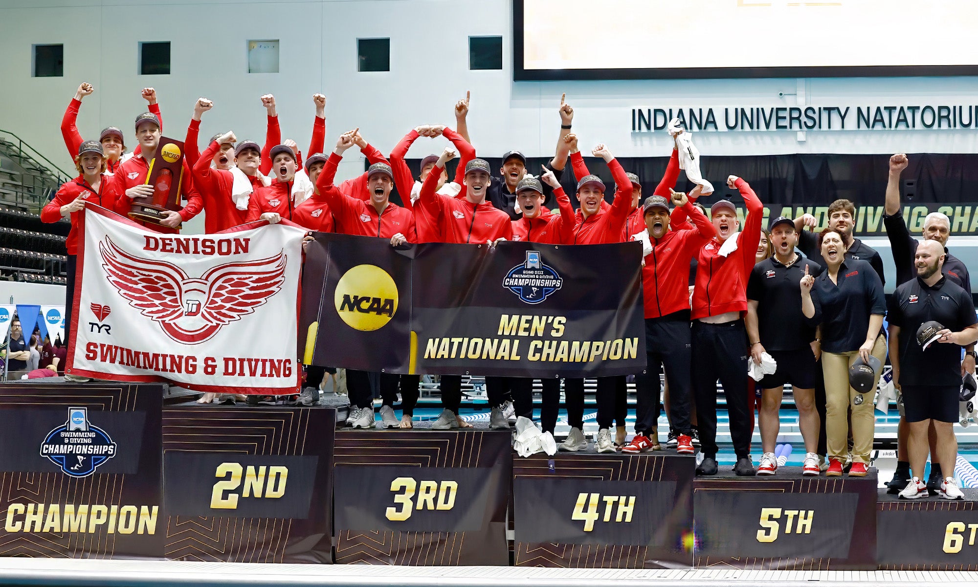 A large group of athletes and coaches in Denison athletics gear stands on a large NCAA podium next to a swimming pool, cheering in celebration.