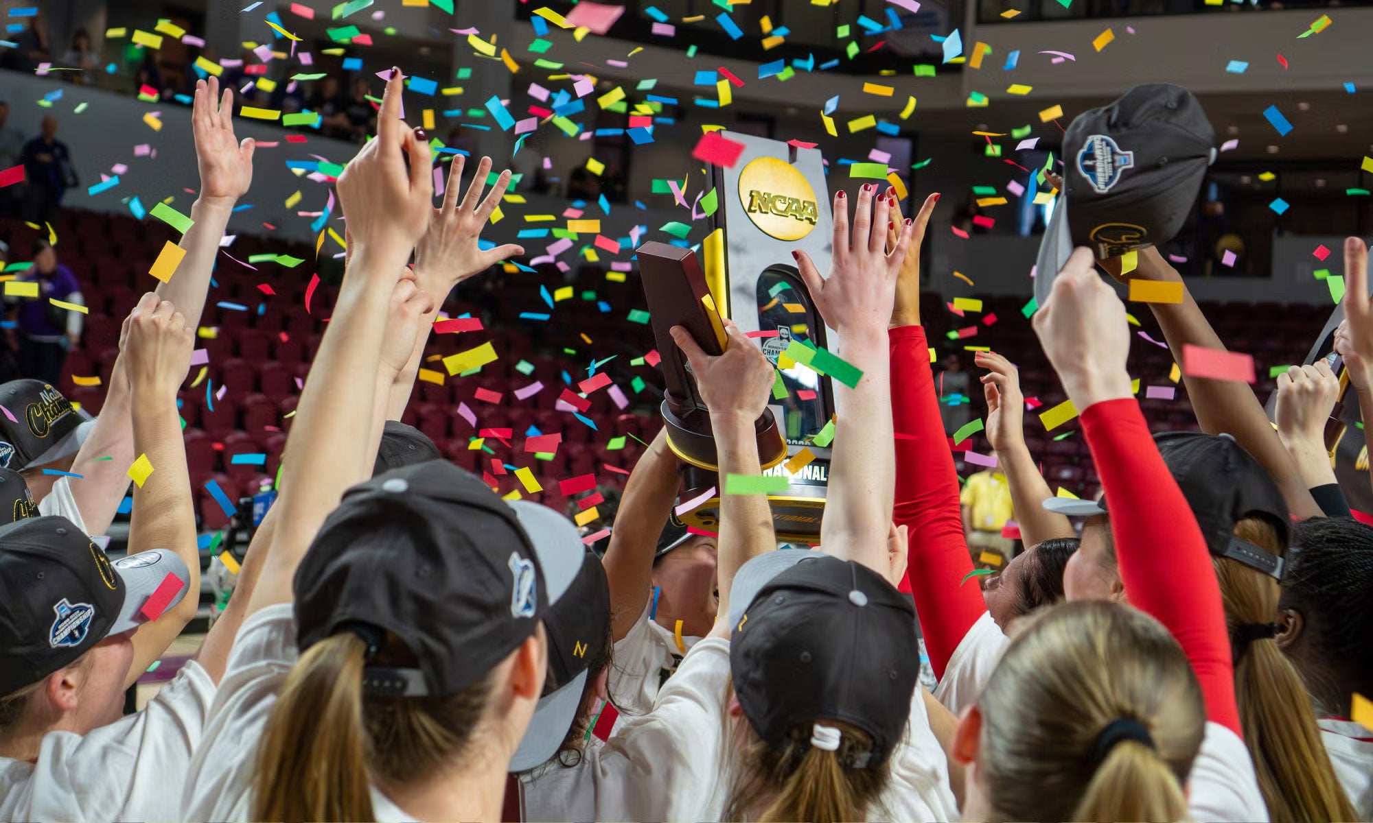 A group of athletes wearing black caps raise their hands and hold a trophy as colorful confetti falls in a crowded indoor arena, celebrating a championship win.