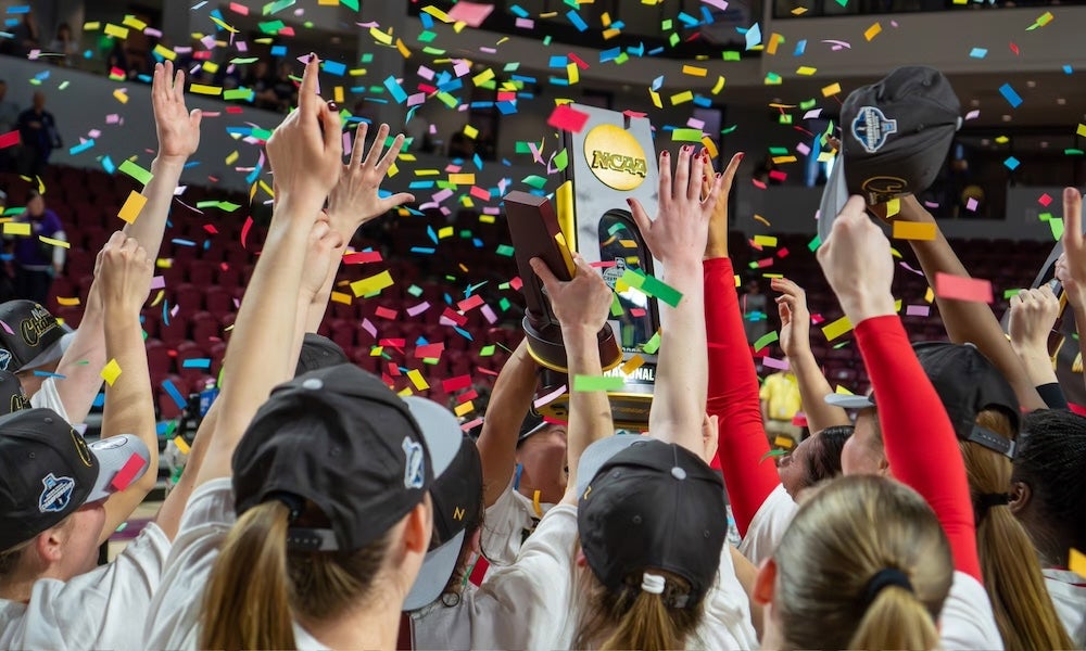 A group of athletes wearing black caps raise their hands and hold a trophy as colorful confetti falls in a crowded indoor arena, celebrating a championship win.