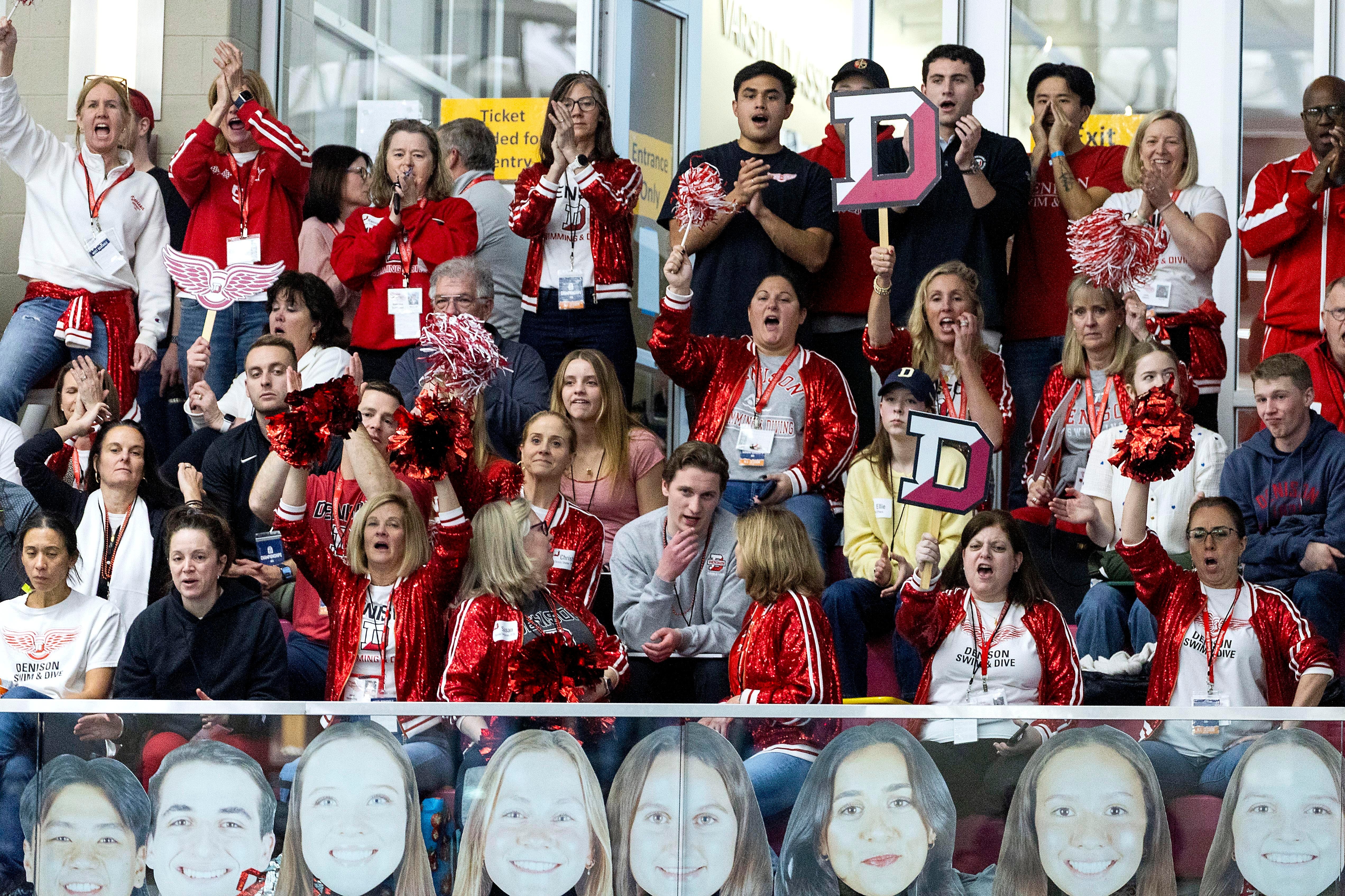A group of fans in red and white clothes cheer from the stands at an indoor athletic event; large cut-out posters of athlete's faces line the first row of the stands..