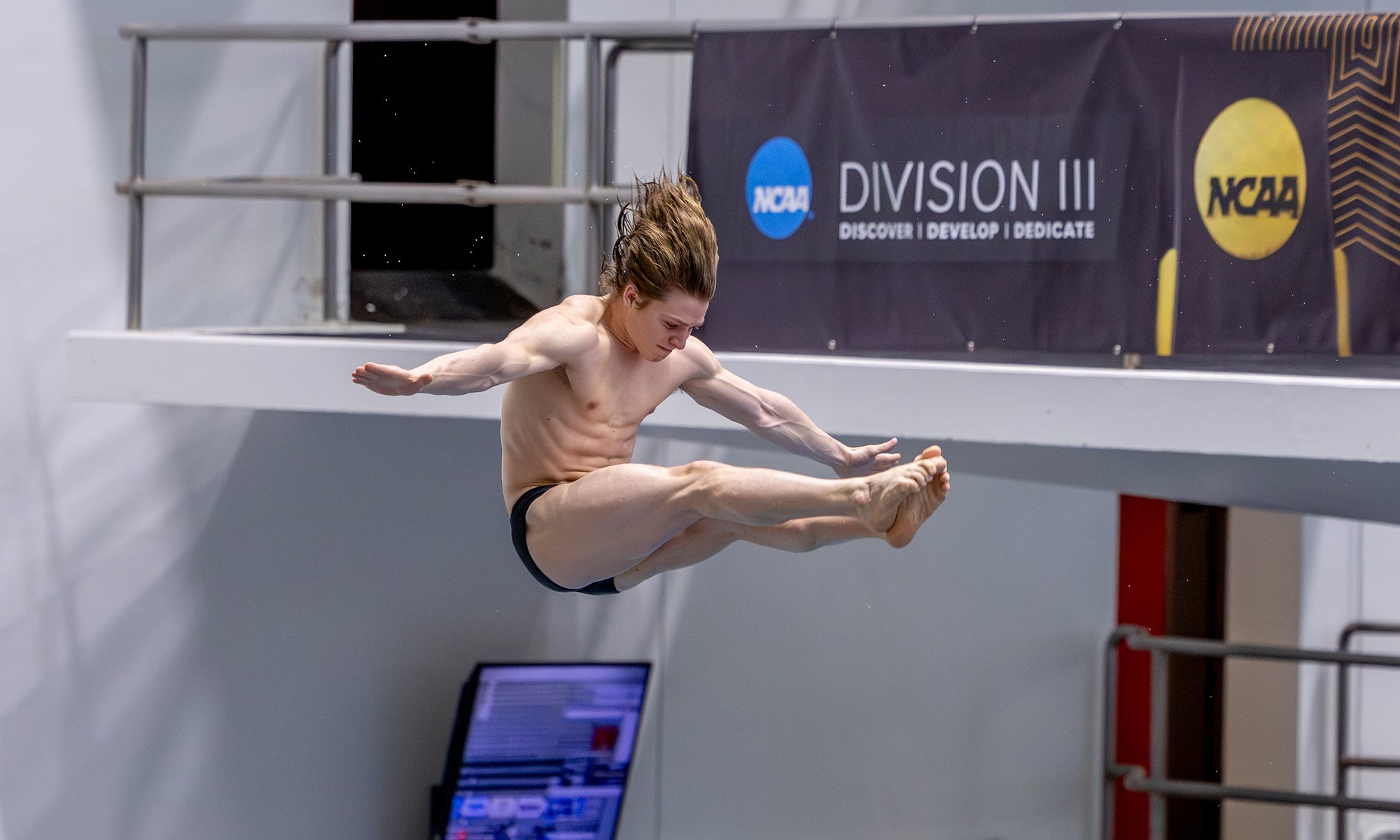 A male diver in mid-air performs a pike position during a dive at an NCAA Division III competition, with a scoreboard and competition banners visible in the background.