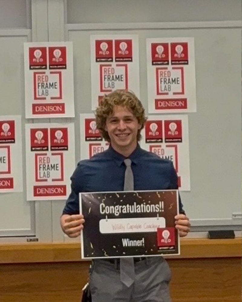 A young man with curly hair, wearing a blue shirt and gray tie, stands indoors holding a sign that says “Congratulations!! Winner!” in front of a board covered with Red Frame Lab posters.