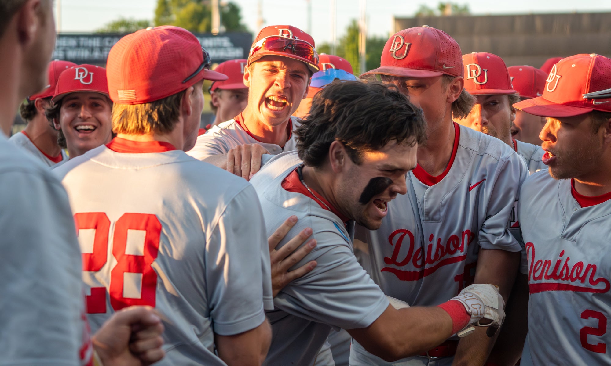 A group of baseball players in gray and red uniforms celebrate together on a field. One player in the center has black eye paint and is being embraced by teammates. The mood appears excited and joyful.