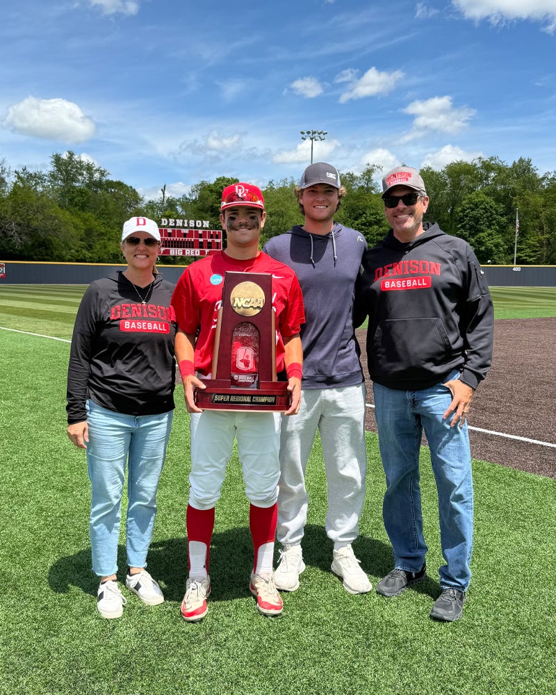 Four people stand on a baseball field posing for a photo. The person second from the left, in a red baseball uniform, holds a large trophy. The other three people wear casual clothes and sunglasses. A scoreboard is visible in the background.