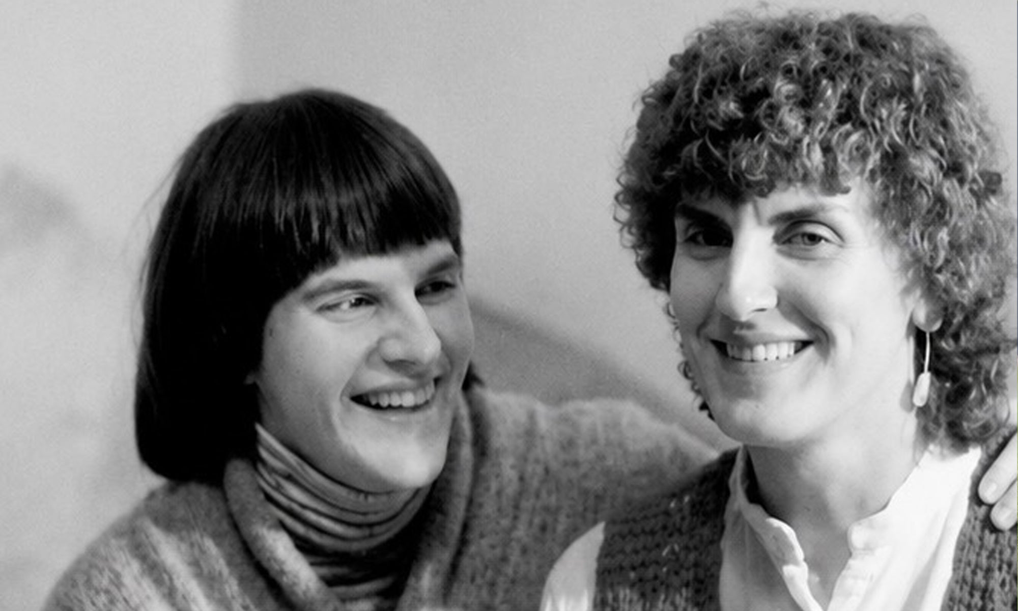 Two women are sitting close together and smiling. One has straight, dark hair with bangs, and the other has curly hair and is wearing earrings. The image is in black and white.
