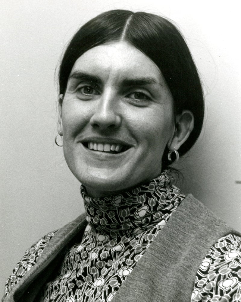 Black and white portrait of a woman with dark hair parted in the middle, wearing a patterned turtleneck and a vest, smiling at the camera against a plain background.
