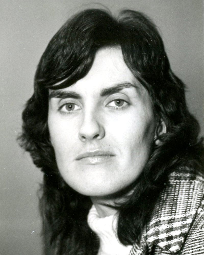 Black-and-white portrait of a person with medium-length dark hair, looking directly at the camera with a neutral expression. The person is wearing a plaid-patterned jacket over a light-colored top.
